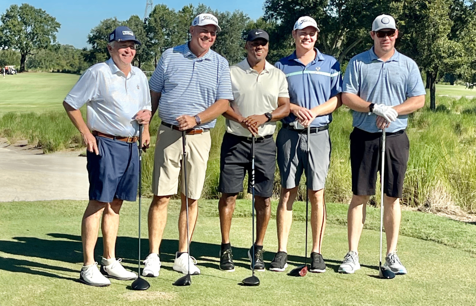 Five golfers lined up at the tee box on a sunny course.